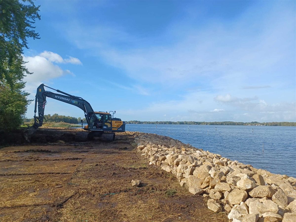 Een strook lichte keien langs het water, met een graafmachine in de achtergrond